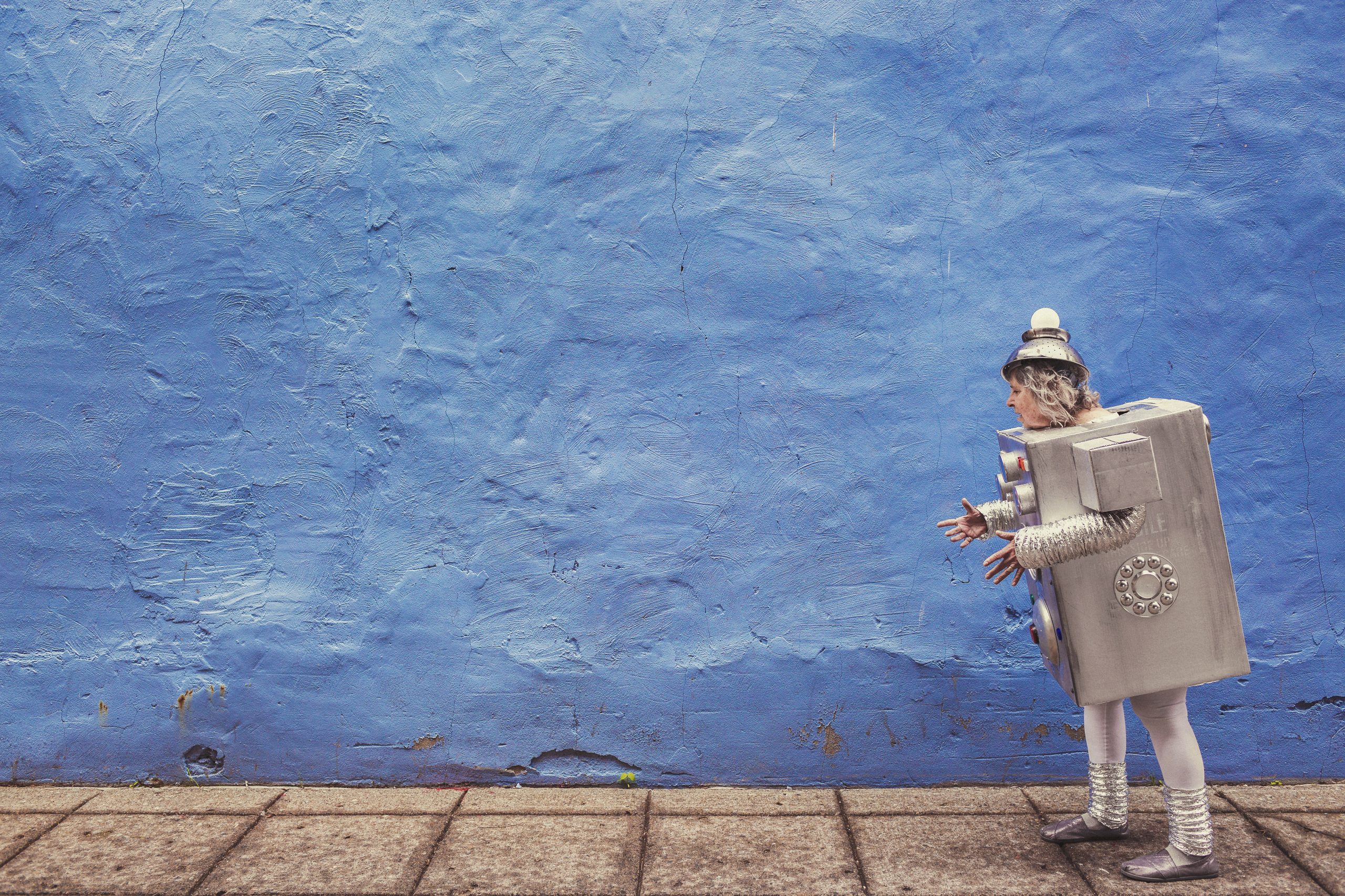 Old women dresses as a silver robot walking past a blue wall.